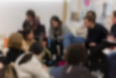 A few people sit in line. Two of them are leaning forward, supporting their elbows on their knees. They lean towards the people who are sitting on the floor, facing them. One of the people on the floor is gesturing with their hands. The other people look towards them.