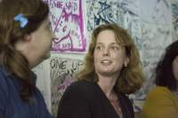 Two people sit next to each other, facing each other. Behind them, there is a wall with drawings and hand-drawn posters.