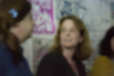 Two people sit next to each other, facing each other. Behind them, there is a wall with drawings and hand-drawn posters.