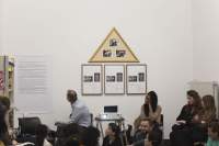 In an indoor space, there are three rectangular frames hanging next to each other, each with two photographs and some text in black. Right on top, there is a yellow triangular frame with three photographs. Below them, there is a low table with a projector, whose light shines towards the camera. One person sits to the left and three people to the right of the projector. The person on the left looks behind, towards the left, to a large white poster with black text, which hangs on the wall. In front of them, there are other people, whose upper bodies are partially visible.