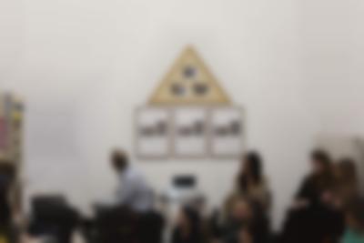 In an indoor space, there are three rectangular frames hanging next to each other, each with two photographs and some text in black. Right on top, there is a yellow triangular frame with three photographs. Below them, there is a low table with a projector, whose light shines towards the camera. One person sits to the left and three people to the right of the projector. The person on the left looks behind, towards the left, to a large white poster with black text, which hangs on the wall. In front of them, there are other people, whose upper bodies are partially visible.