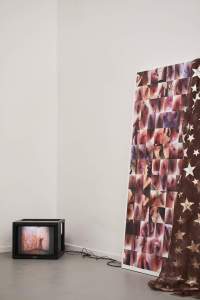 In the corner of a room, a TV set lies on the floor, showing an image. To its right, there is a mixed-media piece, made from a large canvas with several pictures, and a piece of brown fabric with golden stars, which is draped on the right side.