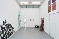 Indoor space with white walls and grey floor. Around the room, there is a frame, a photograph installation, an installation of four pieces of textile, a large blue poster, a mirror, another mixed-media installation and a TV screen. These are all hanging or propped against the walls, except for the TV which is on the floor, in one of the corners.