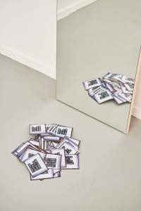 On the floor, several copies of the same image are piled up. A mirror is propped against the wall, showing the reflection of the stack of images.