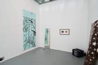Indoor space with white walls and grey floor. Around the room, there is a large blue screen, a mirror, a frame, a TV set and a mixed media installation. These are all hanging or propped against the walls, except for the TV which is on the floor, in the right corner. On the large blue screen there is a hand-drawn horse with rounded black text at the top, reading: 