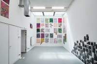 Indoor space with white walls and grey floor. At the back of the room, filling one of the walls, there are several hand-drawn posters. On the wall on the right-hand side, there is an installation made of several black-and-white photographs. Another two pieces are seen hanging on the opposite wall.