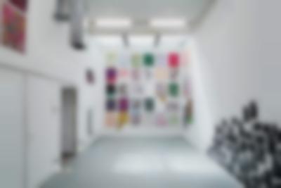 Indoor space with white walls and grey floor. At the back of the room, filling one of the walls, there are several hand-drawn posters. On the wall on the right-hand side, there is an installation made of several black-and-white photographs. Another two pieces are seen hanging on the opposite wall.