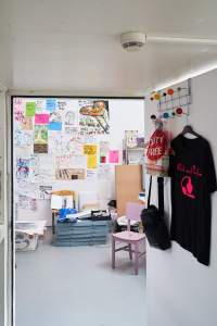 On the right, hanging from an over the door hook, there is a t-shirt, a plastic bag and a handbag. Through the open door, a room is partially visible. There are hand-drawn posters and drawings hanging on the walls, a pink chair, shelfs and other boxes and materials on the floor.