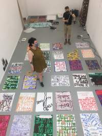 Seen from above, several hand-drawn posters have been set on a grey floor. Two people stand, in different spots, in between the posters. At the back of the room there are other objects, namely an open laptop on the floor.