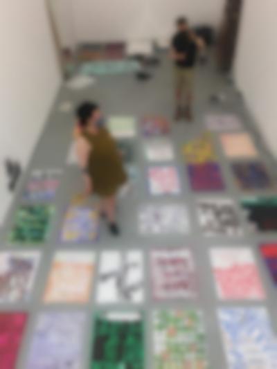 Seen from above, several hand-drawn posters have been set on a grey floor. Two people stand, in different spots, in between the posters. At the back of the room there are other objects, namely an open laptop on the floor.