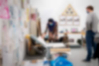 A person is leaning towards an open box with large sheets of paper, which they are handling. Another person, to the right, observes. At the back of the room, there is an desk with an open laptop and frames hanging on the wall. On the left, posters and drawings hang on the wall. There is also a shelf next to this wall, as well as other materials on the floor.