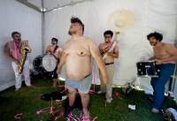 A band is playing inside a white canopy tent. There are five elements in the band, all bare chested. The person at the front, wears only boxers.