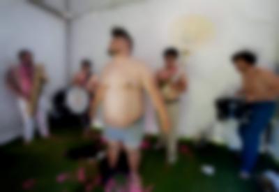 A band is playing inside a white canopy tent. There are five elements in the band, all bare chested. The person at the front, wears only boxers.