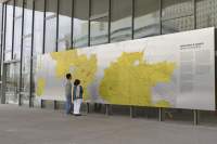 A large mural is outside a building with a glass facade. Two people look at the mural, which has a large map with large yellow areas. A section with a few paragraphs of text is on the right edge of the mural.