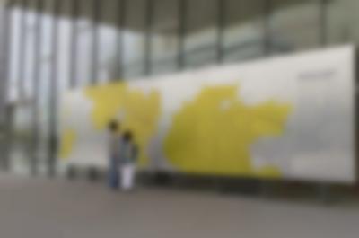 A large mural is outside a building with a glass facade. Two people look at the mural, which has a large map with large yellow areas. A section with a few paragraphs of text is on the right edge of the mural.