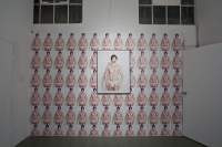 On a white wall of an indoor space a single image appears several times, filling nearly the entirety of a wall. The image appears larger at the centre of the wall, inside a frame. It then appears smaller also on the wall, in six rows. The image shows an American shot of a person wearing only white briefs. They have short hair and their lips are painted red.