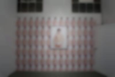 On a white wall of an indoor space a single image appears several times, filling nearly the entirety of a wall. The image appears larger at the centre of the wall, inside a frame. It then appears smaller also on the wall, in six rows. The image shows an American shot of a person wearing only white briefs. They have short hair and their lips are painted red.