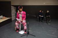 A person sits behind a microphone. They are wearing a bright pink blazer and high heels, a lighter pink scarf and headgear and trousers and a top with pink hues. They are holding paper with their hands. There are also two sheets of paper on the floor, in between their feet. At the back, two other people sit behind another microphone.