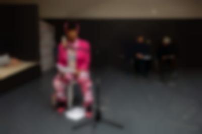 A person sits behind a microphone. They are wearing a bright pink blazer and high heels, a lighter pink scarf and headgear and trousers and a top with pink hues. They are holding paper with their hands. There are also two sheets of paper on the floor, in between their feet. At the back, two other people sit behind another microphone.