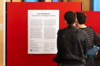 Two people read an exhibition text, which appears inside a white poster on a red wall. At the top, the text reads in larger font 