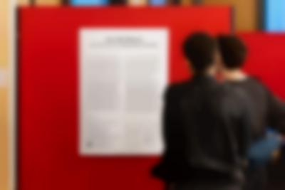 Two people read an exhibition text, which appears inside a white poster on a red wall. At the top, the text reads in larger font 