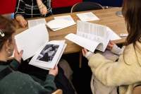 Three people sit around a wooden table. There are booklets and other printouts on the table. The two people with their backs to the camera are leafing through a booklet and a printout stapled at the top left corner. The printout shows the photograph of a person wearing sunglasses.