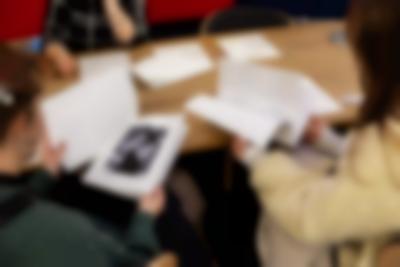 Three people sit around a wooden table. There are booklets and other printouts on the table. The two people with their backs to the camera are leafing through a booklet and a printout stapled at the top left corner. The printout shows the photograph of a person wearing sunglasses.