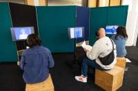 Three people sit in front of three screens, wearing headphones. The screens all show different images of single people.