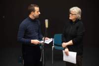 Two people stand next to a microphone. They are both holding paper and looking at each other.