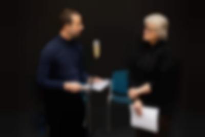 Two people stand next to a microphone. They are both holding paper and looking at each other.