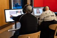 Two people sit in front of screens. They both have headphones on. The person on the left touches the screen, in which there is a picture of a person and a purple block with one word inside. On the right screen, an image of a person wearing a moustache and headgear is visible.