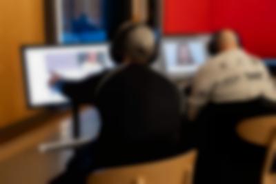 Two people sit in front of screens. They both have headphones on. The person on the left touches the screen, in which there is a picture of a person and a purple block with one word inside. On the right screen, an image of a person wearing a moustache and headgear is visible.