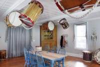 Indoor space with wooden floors, a small window and a table. There are 7 blue chairs around the table. Several large bass drums, or bombos, are hanging from the ceiling. There are smaller ones on the table and by the window.
