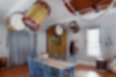 Indoor space with wooden floors, a small window and a table. There are 7 blue chairs around the table. Several large bass drums, or bombos, are hanging from the ceiling. There are smaller ones on the table and by the window.