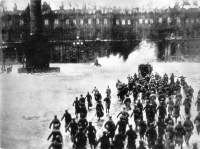 Black-and-white photograph shows a large group of people walking in the same direction in an open field. There is a large building in the background.