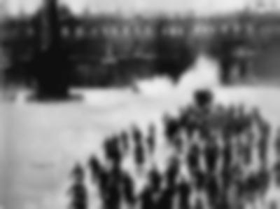 Black-and-white photograph shows a large group of people walking in the same direction in an open field. There is a large building in the background.