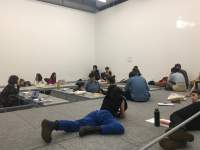 A group of people sits around low tables or on the floor, which is on a higher level, on par with the tables. One of the people lies, belly down, on their elbows and another person lies on their side. People are grouped in twos and are engaging in conversation.