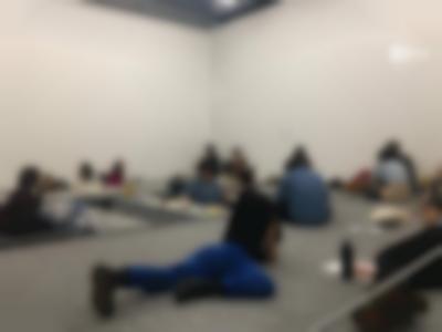 A group of people sits around low tables or on the floor, which is on a higher level, on par with the tables. One of the people lies, belly down, on their elbows and another person lies on their side. People are grouped in twos and are engaging in conversation.