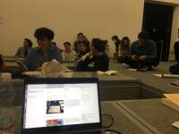 An open laptop screen shows an open website with a white background, two images and some text. Behind it, a group of people sits around low tables or on the floor, which is on a higher level, on par with the tables.