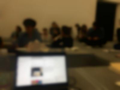 An open laptop screen shows an open website with a white background, two images and some text. Behind it, a group of people sits around low tables or on the floor, which is on a higher level, on par with the tables.