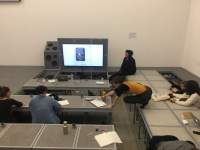 A group of people sits around low tables. At the front, a screen shows a projected image. Someone sits to the right of the screen. One of the people is on top of a table, and stretches themselves to reach something on a nearby table.