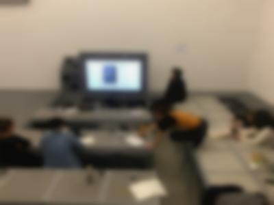 A group of people sits around low tables. At the front, a screen shows a projected image. Someone sits to the right of the screen. One of the people is on top of a table, and stretches themselves to reach something on a nearby table.