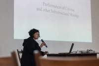 A person sits at a table, with an open laptop. They are holding a microphone close to their face. Behind them, a large slide is being projected on the wall. The slide reads 