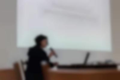 A person sits at a table, with an open laptop. They are holding a microphone close to their face. Behind them, a large slide is being projected on the wall. The slide reads 