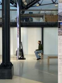 In a lofty indoor space, a person sits in front of a screen, which hangs from the ceiling. There is a monitor in front of the screen, which they look towards. There is a black column in the space, visible at the front, and a mezzanine level, visible at the back.