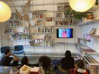 Five people sit on the floor, looking up at a small screen with the image of a person with long hair, dressed in a red dress. The screen is hung to the wall, and there are several shelfs with books around it.