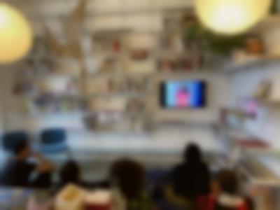 Five people sit on the floor, looking up at a small screen with the image of a person with long hair, dressed in a red dress. The screen is hung to the wall, and there are several shelfs with books around it.