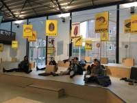 Indoor space with large windows. There is a stage-like structure with two levels. There are several signs on poles, set on this structure. They have yellow backgrounds, an illustration and a word underneath. The words appear in different languages and in the illustrations several animals appear: a rat, fishes, a turtle, etc. There are six people sitting on the first level.