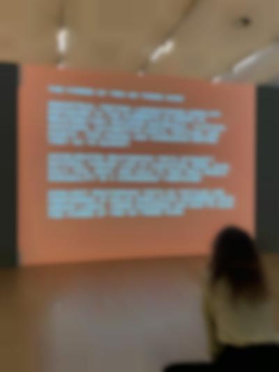 A person is sitting in front of a large screen. On the screen, a text in white is being projected.