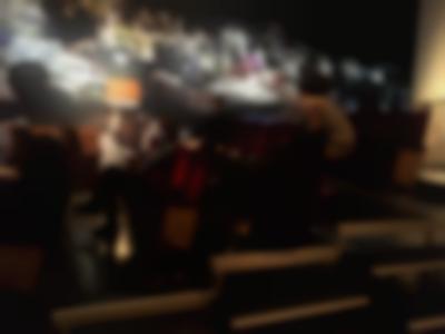 Several people, wearing face masks, sit in four rows of tribute sitting. Many seats are left empty. There is a floor lamp at the back. The only visible wall has a large black-and-white photograph as wallpaper.